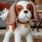 A crocheted toy dog designed to resemble a cute, small breed with white and brown coloring, featuring large button-like eyes and a collar with a medallion, standing on a wooden floor in a cozy living room setting.