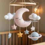 A baby mobile hanging over a crib, featuring crocheted elements including a crescent moon, clouds, stars, and a ringed planet in pastel colors.