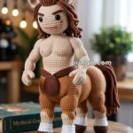 A crocheted centaur figure with a human upper body and a horse lower body, featuring brown and beige yarn, standing on a wooden table with a book titled 'Mythical Creatures' in the background.