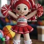 A crocheted doll dressed as a cheerleader with red and white pom-poms, featuring the text 'TEAM SPIRIT' on her top. She stands on an orange and white crocheted mat, with a playful and colorful background, including a basketball.
