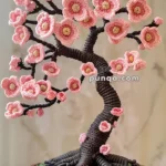 A crochet art piece depicting a cherry blossom tree with pink flowers, placed in a decorative pot with floral designs.