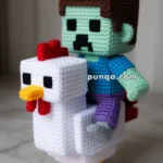 A crocheted doll resembling a pixelated character riding a white chicken, both made with blocky features reminiscent of a popular video game style.