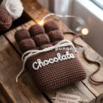 A brown knitted item resembling a chocolate bar with the word "Chocolate" stitched on it, tied with a white string, resting on a wooden surface. Nearby, there is a label reading "handmade with love" and balls of yarn in the background.