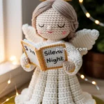 A handmade, crocheted angel figure wearing a white robe and holding a book labeled 'Silent Night', with a yellow halo above its head, set against a softly-lit background adorned with fairy lights.