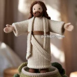 A crocheted Christ The Redeemer Statue doll with long brown hair and a beard, wearing a long cream robe with a brown belt, stands atop a similarly crocheted hill with green patches. The scene appears homemade with a blurred, neutral background.