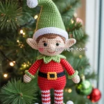 Crocheted elf ornament in red and green, hanging on a Christmas tree with lights and baubles in the background.
