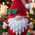 A crocheted Christmas gnome with a red hat and white beard, surrounded by decorative items like a small gift, a tiny Christmas tree, and holly leaves, set against a festive background with candles and blurred Christmas lights.