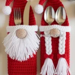Two crocheted Santa-themed utensil holders, one with a white beard and the other with white braids, holding silverware against a festive background with pine branches, pinecones, and jingle bells.