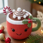 A crocheted red mug with a smiling face, decorated with whipped cream, marshmallows, and a candy cane, sits on a wooden table amidst Christmas decorations.