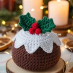 A crocheted holiday pudding with white icing, green holly leaves, and red berries, placed on a wooden coaster, surrounded by cinnamon sticks and festive lights, with candles in the background.