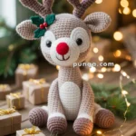 A crocheted reindeer toy with a red nose and a green holly decoration sits on a wooden table surrounded by small, gift-wrapped boxes and festive lights, with a blurred Christmas tree in the background.