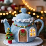 A cozy crocheted teapot designed as a snow-covered house, complete with a wreath on the door and lit windows, sitting on a tabletop surrounded by snowflake decorations.