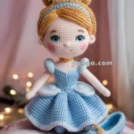 A crocheted doll dressed in a blue gown with blonde hair styled in a bun and a small tiara, standing on a pink cushion with a crocheted blue shoe next to it.