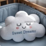 A white cloud-shaped pillow with eyes closed and a smile, featuring the words "Sweet Dreams" embroidered on it, placed on a crib with a light blue mattress.