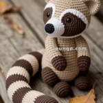 A handmade crocheted raccoon toy sitting on a wooden surface with an autumn leaf nearby.