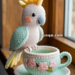 A crocheted bird with pastel colors perched on the rim of a crocheted teacup, which has floral designs, sitting on a windowsill.