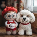 Crochet dolls, one wearing a red beret and glasses holding a coffee mug, and the other resembling a fluffy white dog with a red collar, positioned on a wooden surface by a window with lace curtains.