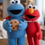 Two crocheted plush toys, one blue holding a brown cookie, and one red with a large orange nose, standing on a wooden surface in a cozy room setting.