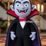 A crocheted vampire doll with pointed ears, green eyes, and sharp fangs, dressed in a black suit with a red bow tie and a cape, standing in a room with lit candles in the background.