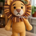 A crocheted amigurumi lion with a fluffy mane, featuring a small green bow on its head, standing on a wooden table in a softly lit room.