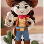 Crocheted doll dressed as a cowgirl with a brown hat, red scarf, and holding a lasso, standing on a woven mat with small crocheted cacti and rocks.