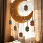 A crocheted yellow crescent moon mobile hanging by a window, adorned with stars and colored spheres. Below, an open book rests on a stand, with curtains filtering sunlight into the room.