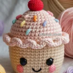 A crocheted cupcake toy with a smiling face, featuring a pink icing top, colorful sprinkles, and a red cherry on top, surrounded by balls of yarn.
