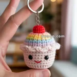 A hand holding a keychain featuring a crocheted cute cupcake with a rainbow top and a smiling face, displayed against a blurred indoor background.