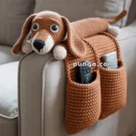 A crocheted brown dachshund armrest organizer draped over the arm of a sofa, featuring two pockets holding a remote control and a smartphone.