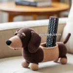 A crocheted toy shaped like a brown and beige dachshund, designed to hold a remote control, placed on a beige sofa.