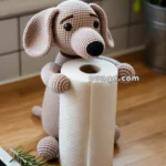 A crocheted dog holding a roll of paper towels on a wooden kitchen counter, with a sprig of rosemary next to it.