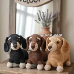 Three crocheted dachshund plushies in black, brown, and beige sit on a wooden table in front of a mirror with a "welcome" sign.