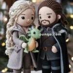 Two crocheted dolls dressed in medieval-style clothing stand together on a wooden platform, each wearing detailed outfits with textured patterns. The doll on the left holds a small green and orange crocheted dragon. Both dolls have intricate hairstyles and facial features, set against a blurred background with warm, glowing lights.
