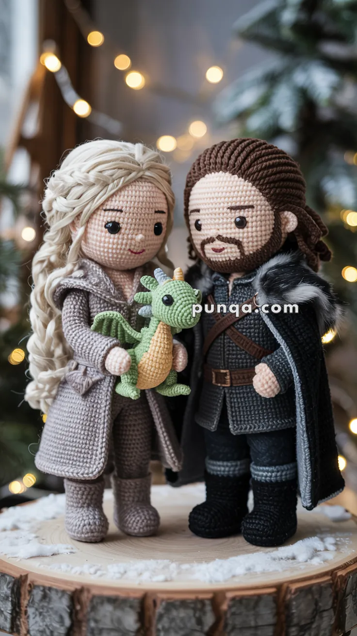 Two crocheted dolls dressed in medieval-style clothing stand together on a wooden platform, each wearing detailed outfits with textured patterns. The doll on the left holds a small green and orange crocheted dragon. Both dolls have intricate hairstyles and facial features, set against a blurred background with warm, glowing lights.