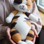 A person holding a crocheted cat toy with a mix of brown, orange, and white colors, featuring a whimsical and smiling face.