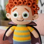 A handmade crocheted doll with curly orange hair, large green eyes, wearing a striped green and yellow shirt, blue pants, and a black cape.