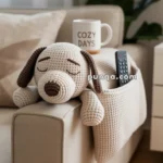 A crocheted plush dog with closed eyes rests on the arm of a beige couch, accompanied by a mug labeled 'Cozy Days' and a remote in a side pocket.