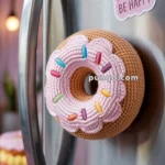 A crocheted doughnut with pink icing and colorful sprinkles is attached to a refrigerator door. Next to it, a sticker reads, 'DONUT WORRY BE HAPPY.' The background features a softly lit kitchen scene.