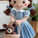 A crocheted doll wearing a blue dress, red shoes, and holding a basket with a small crocheted teddy bear inside.