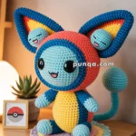 A crocheted doll resembling a cute creature with large, colorful ears, a blue face, and a yellow belly, sitting on a wooden table beside a small plant and a framed Pokeball design.