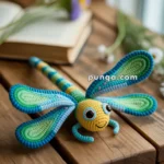A colorful crocheted dragonfly toy with large, expressive eyes rests on a wooden surface, with blurred flowers and an open book in the background.