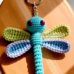 A crocheted dragonfly keychain with a blue body, large purple eyes, and wings in green and blue hues, resting on a wooden surface in sunlight.