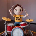 Crocheted doll playing a red and yellow drum set, wearing a yellow shirt with 'Crochet Rock' printed on it, set against a blurred background with hanging lights.