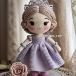 Crocheted doll with a lavender dress and a matching crown, standing on a lace doily next to a rose, in front of a lace curtain.
