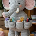 A crocheted elephant figure with multiple pockets on its body, holding various stationery items like scissors and pens, displayed on a wooden table with yarn and crafting tools nearby.