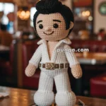 A crocheted Elvis Presley doll in a white jumpsuit stands on a record player labeled "HOUND DOG" in a retro-style setting.