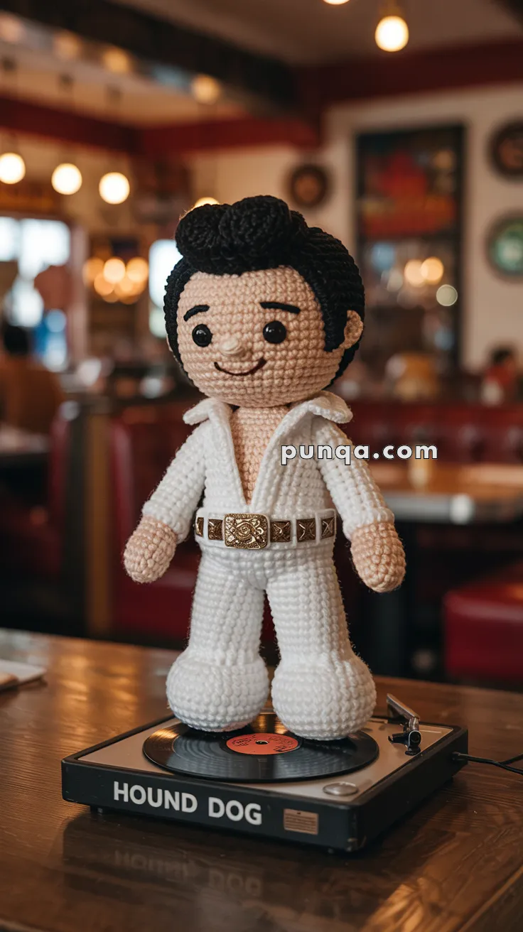 A crocheted Elvis Presley doll in a white jumpsuit stands on a record player labeled "HOUND DOG" in a retro-style setting.