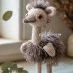 A handmade crochet emu toy with shaggy gray fur and large orange eyes, standing on a wooden surface near a window, with a plant in the foreground.