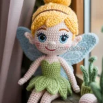 Crocheted fairy doll with yellow hair styled in a bun, wearing a green dress and blue wings, sitting on a wooden surface by a window.