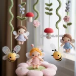 A crocheted baby mobile with fairy and bee characters hanging from a green circular frame, featuring colorful mushrooms, flowers, and a fairy seated on a pink flower base.